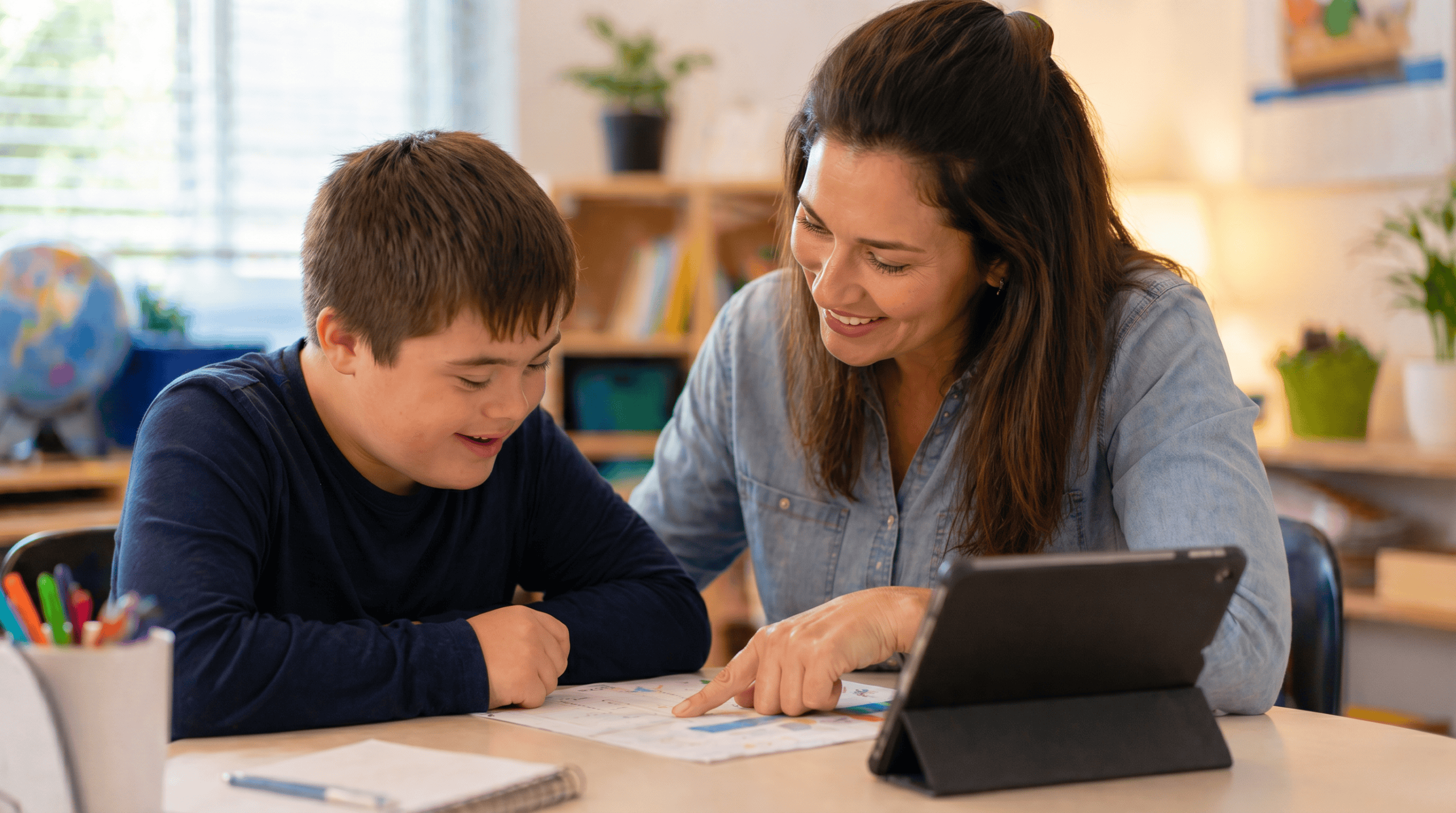 Child with special needs smiling while working on schoolwork at a desk with a supportive tutor nearby