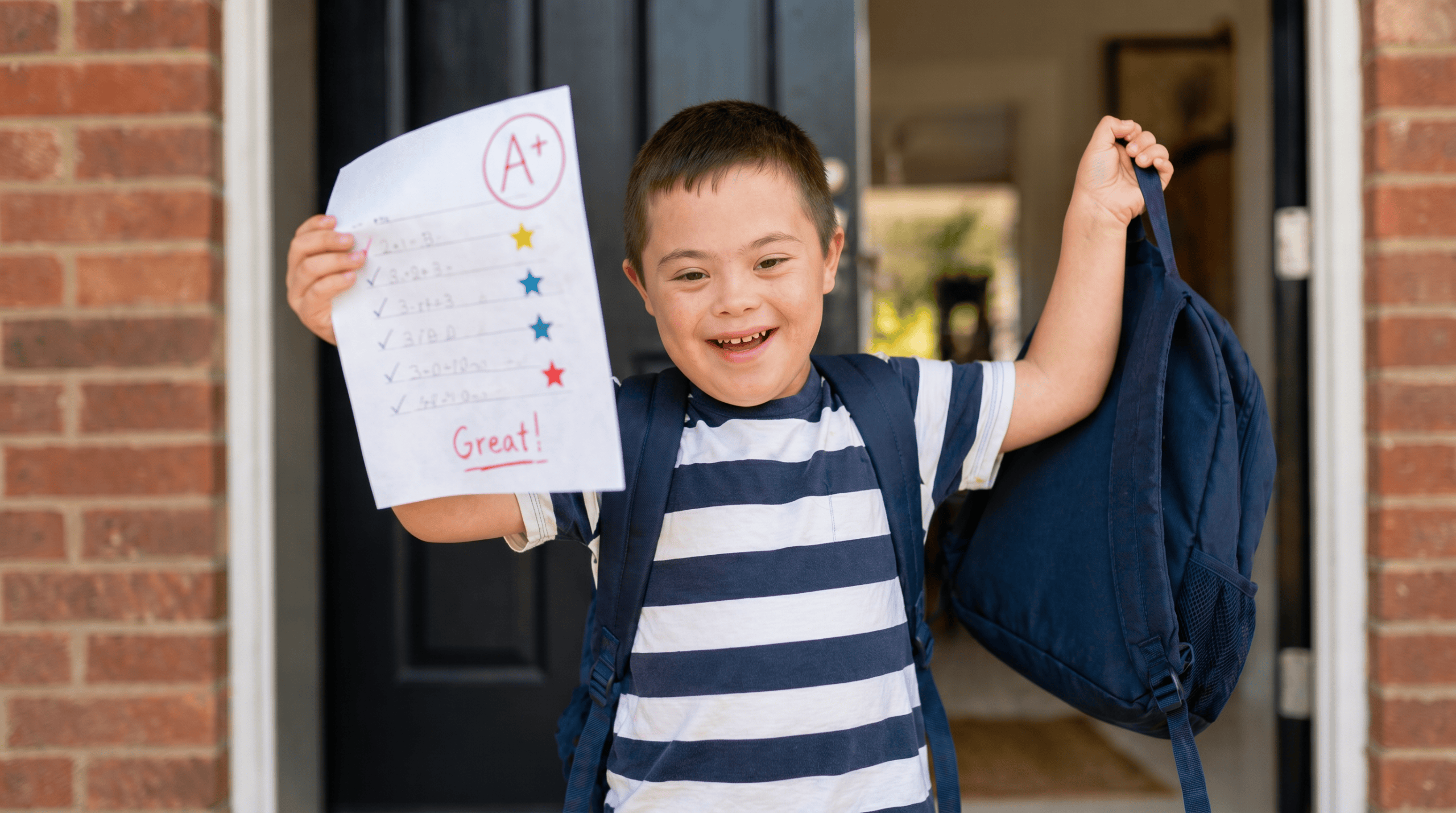 Child with special needs smiling while working on schoolwork at a desk with a supportive tutor nearby